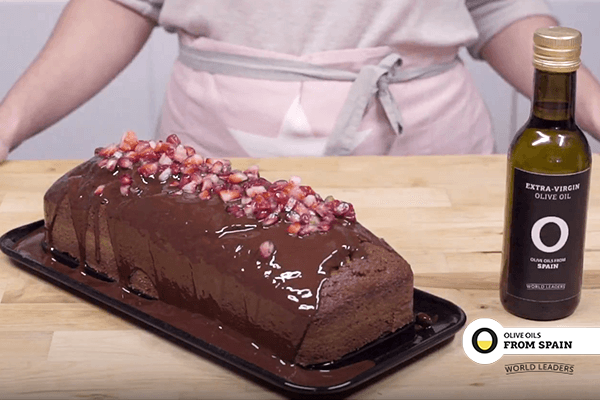 Black tray on wooden table with chocolate sponge covered in melted chocolate decorated with pomegranate. On the table bottle of olive oil. In the background someone with an apron.